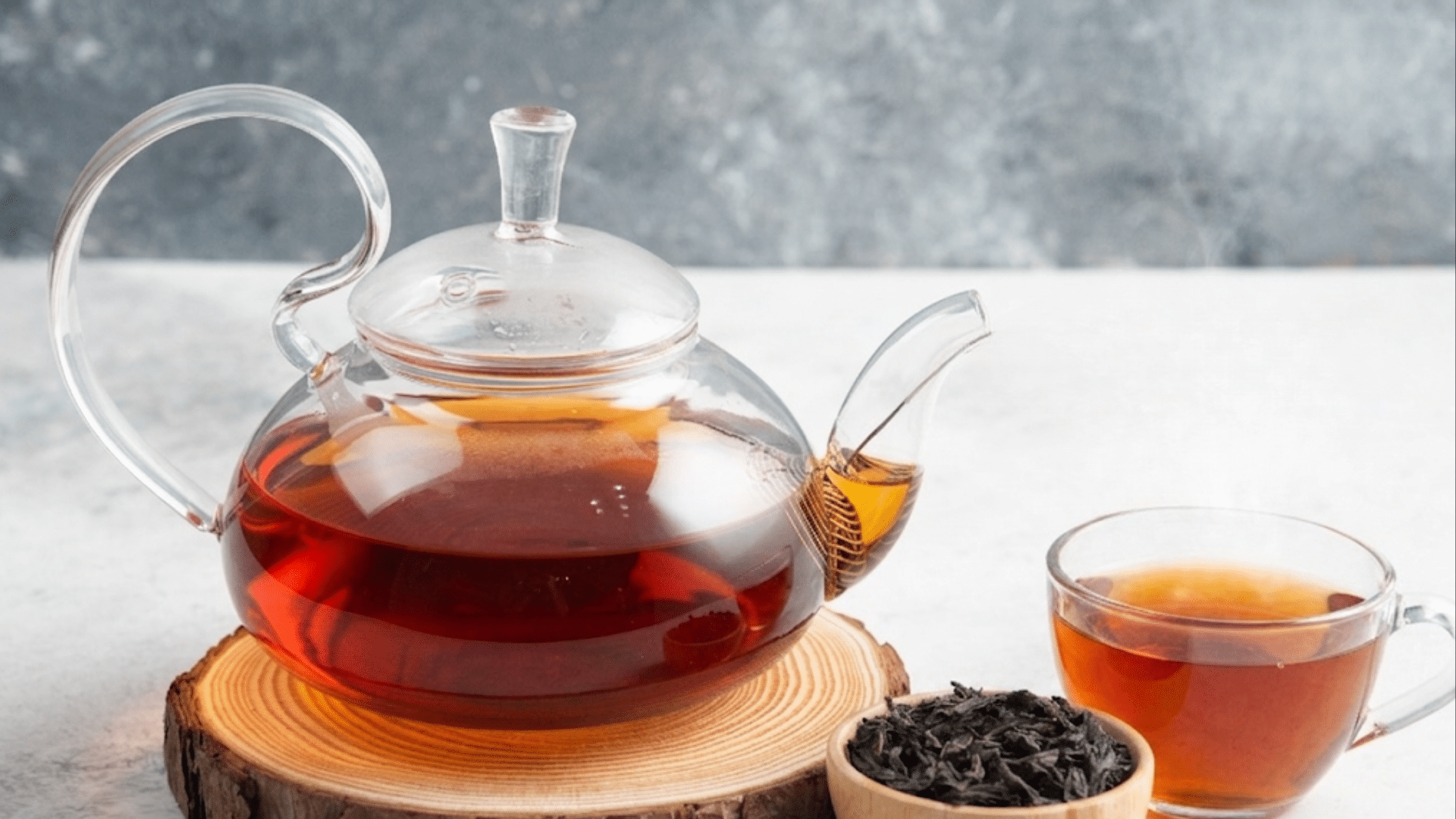 glass teapot filled with freshly brewed earl grey tea beside a steaming glass teacup and loose black tea leaves