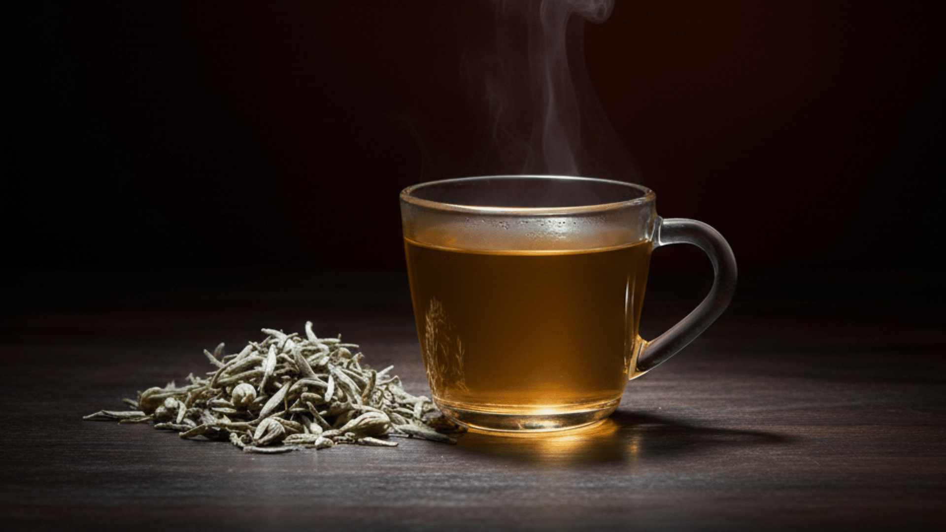 steaming glass cup of hot white tea with a pile of dried white tea leaves on a dark wooden table surface