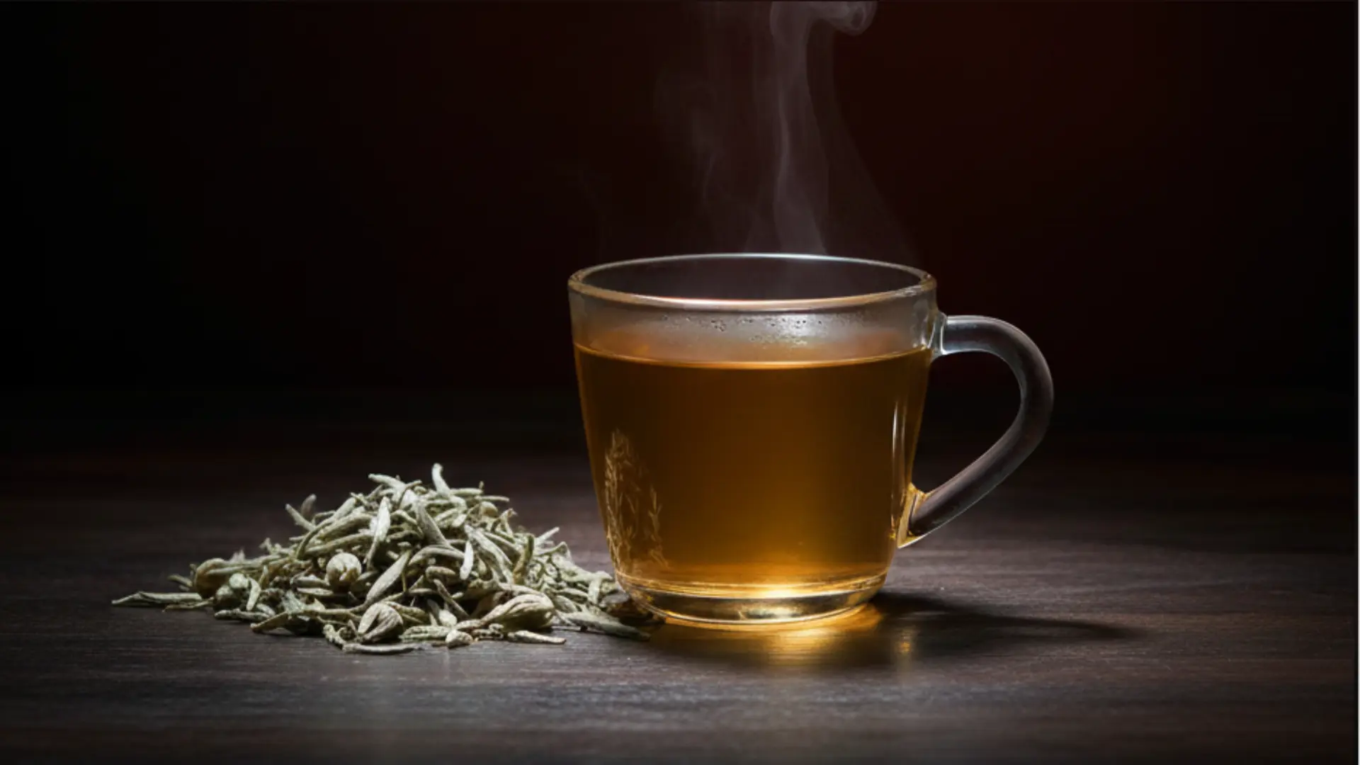 steaming glass mug of brewed tea stands next to a small, neat pile of dried white tea leaves indoors