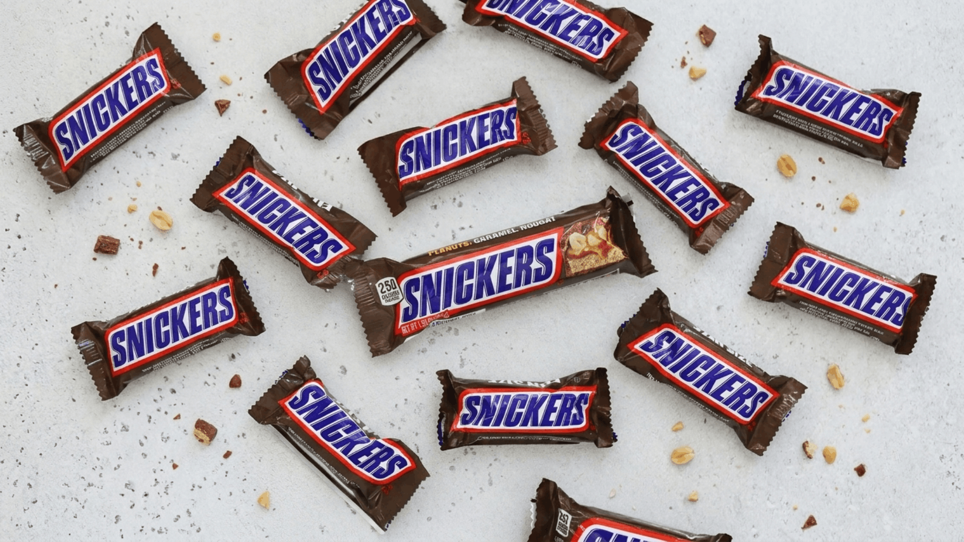 classic snickers candy bars of different sizes with scattered peanuts and crumbs on a grey stone slab