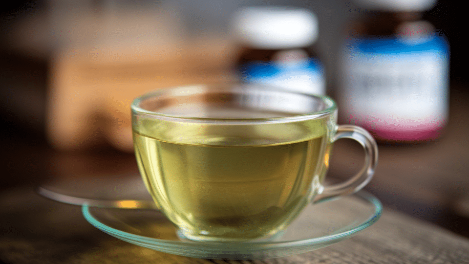 clear glass cup of green tea on a saucer with some supplement bottles on a wooden counter behind the teacup Reply