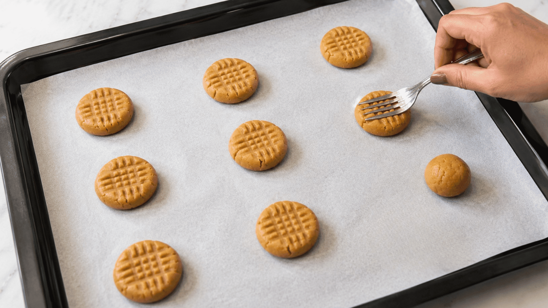 a hand pressing peanut butter cookie dough with a fork creating the classic crisscross pattern on a parchment lined baking sheet