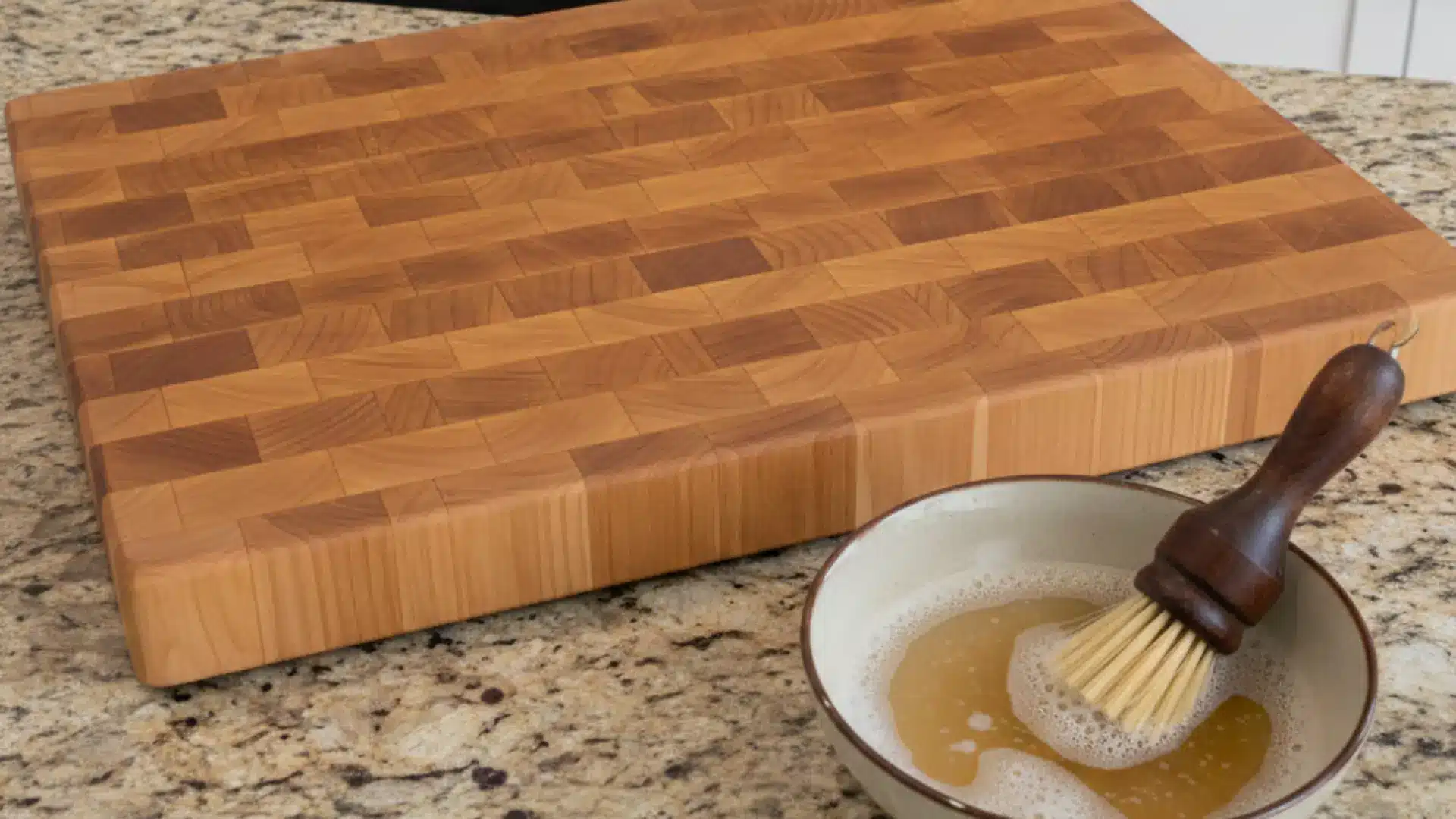 a large wooden cutting board lies flat on a granite kitchen island next to a small bowl of soapy water and a wooden scrub brush