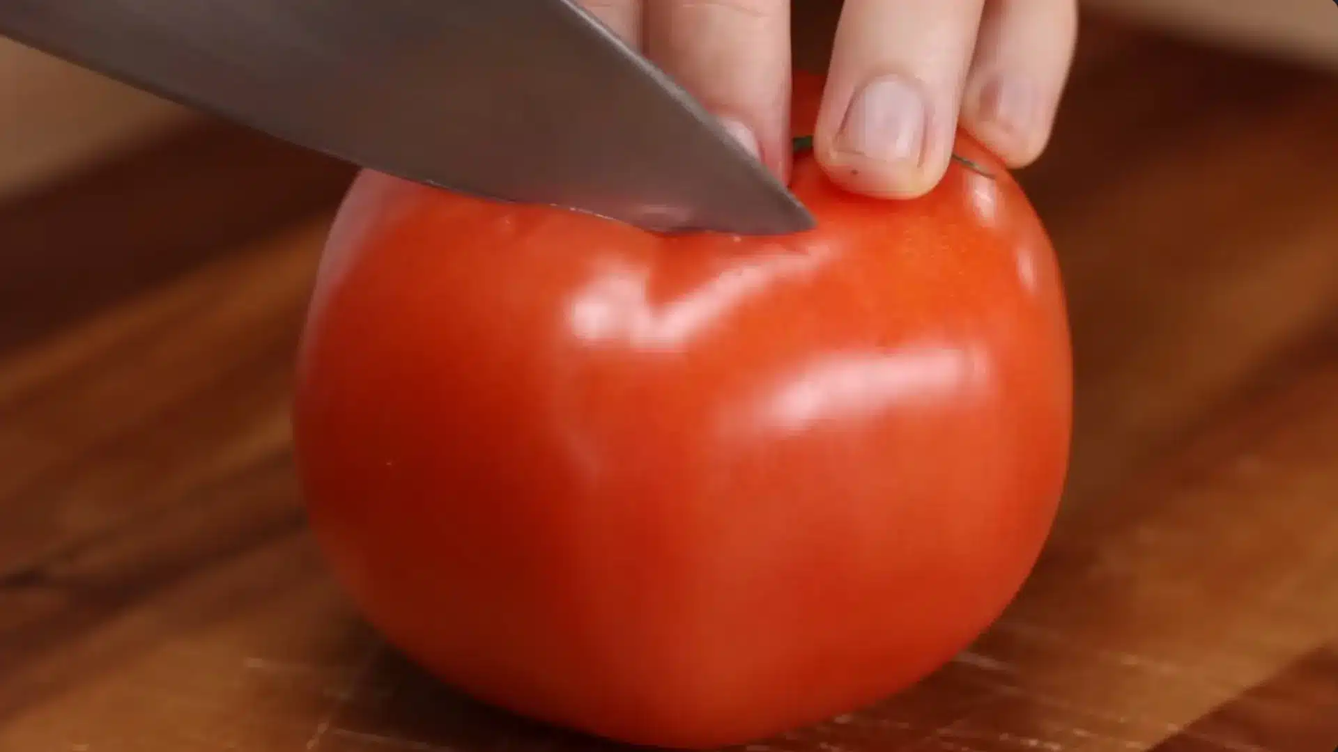 a sharp kitchen knife easily slices through the skin of a fresh red tomato on a wooden cutting board