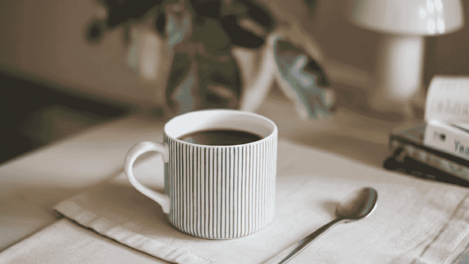 a striped mug with black coffee, sitting on a soft beige napkin, a silver spoon rests next to the mug