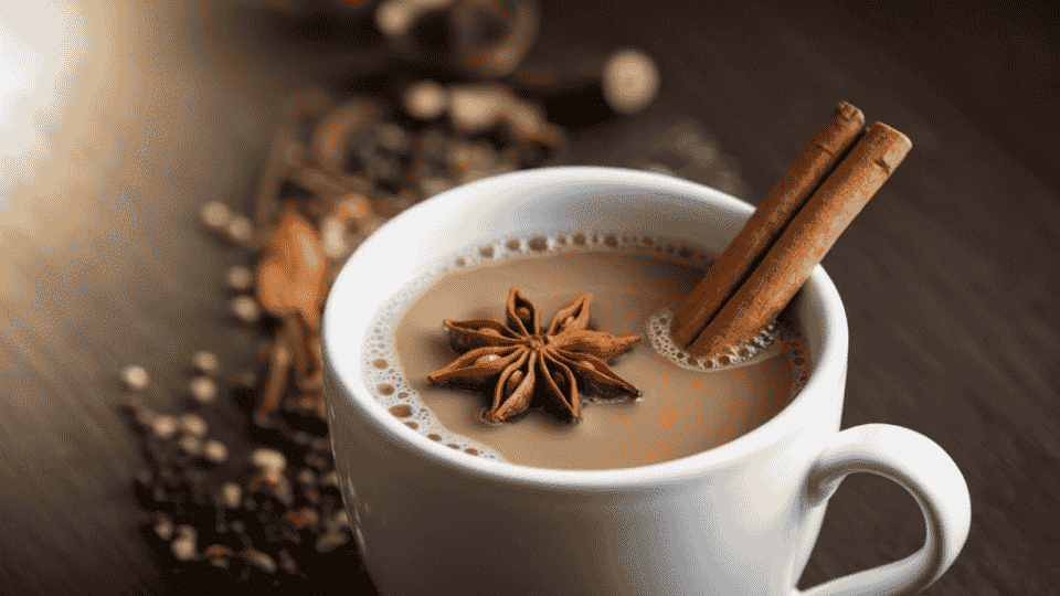 a warm cup of chai with cinnamon sticks and star anise, surrounded by many spices on a wooden surface