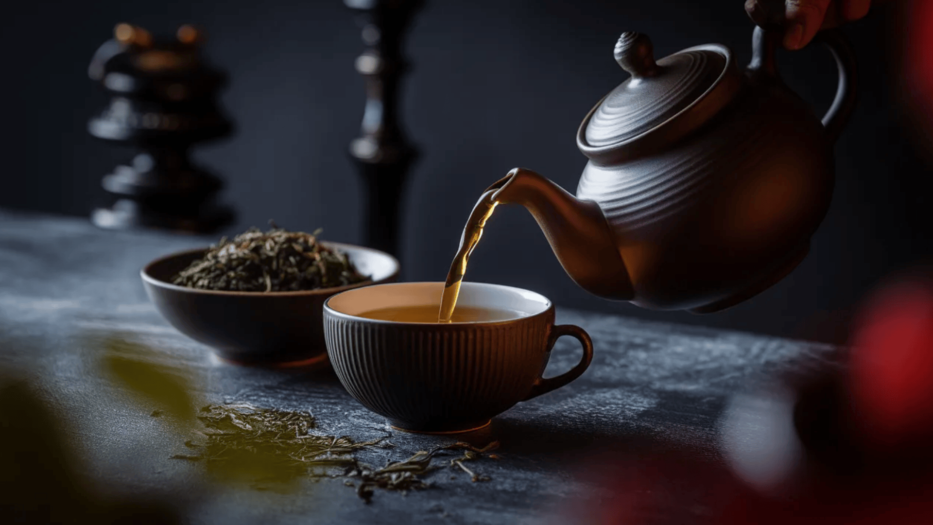 ceramic teapot pouring golden tea into a white cup on a textured surface, with green tea leaves in a bowl and scattered nearby