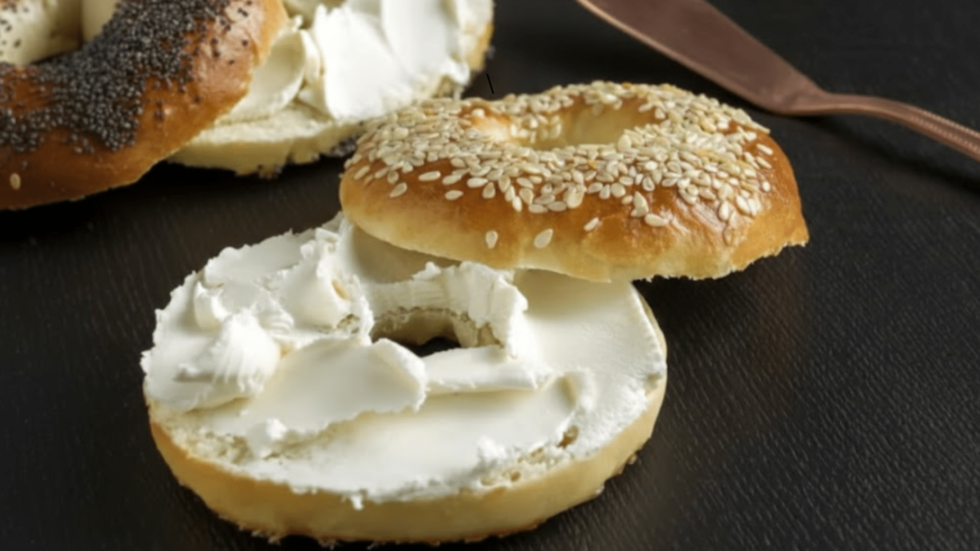 freshly baked sesame and poppy seed bagels sliced and spread with thick, creamy white cream cheese on a dark, textured surface