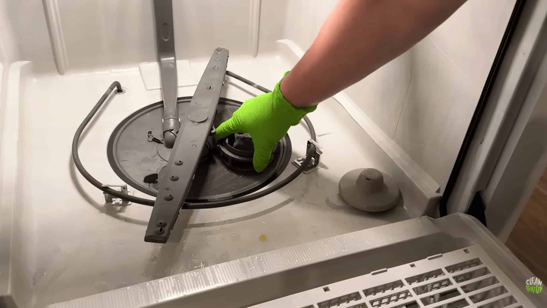 gloved hand removing dishwasher filter from bottom rack area with spray arm and drain visible