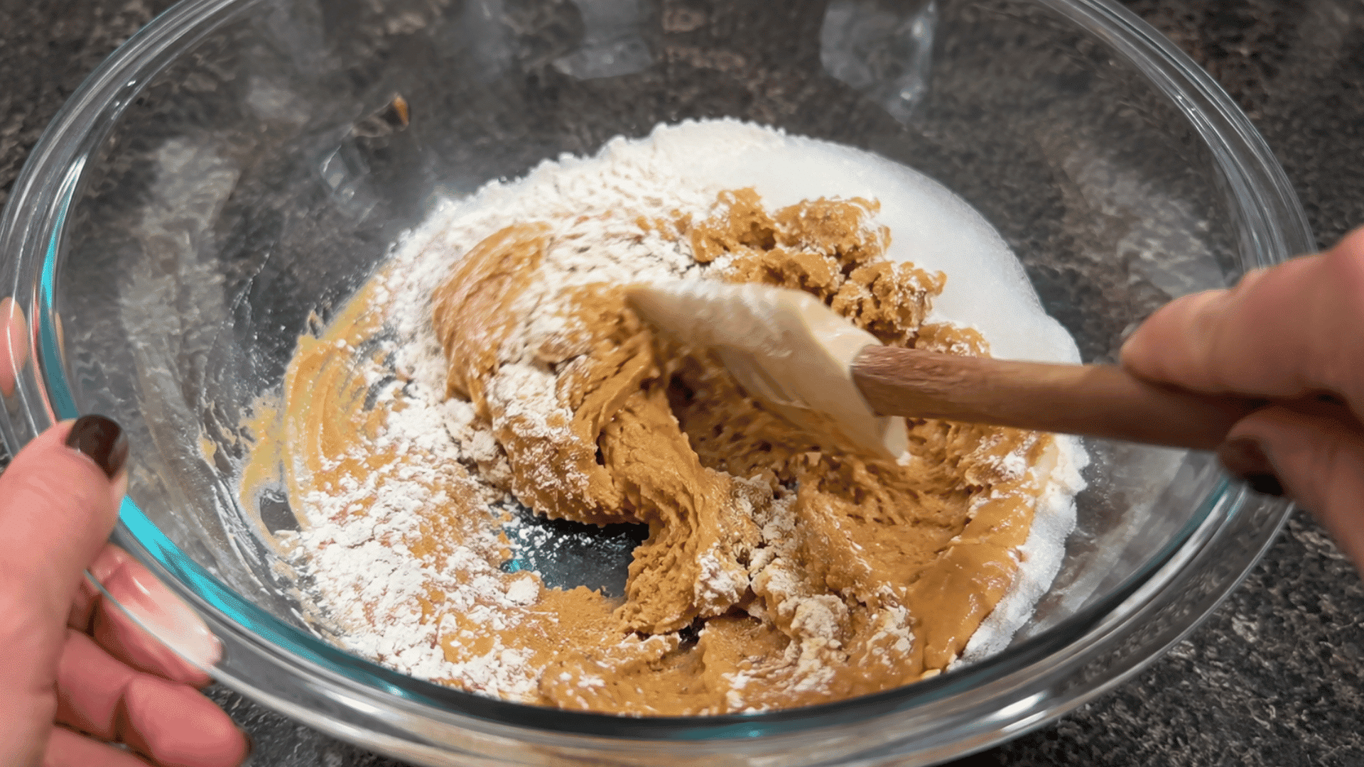 hands mixing peanut butter cookie dough with flour and sugar in a clear glass bowl using a spatula on a kitchen countertop