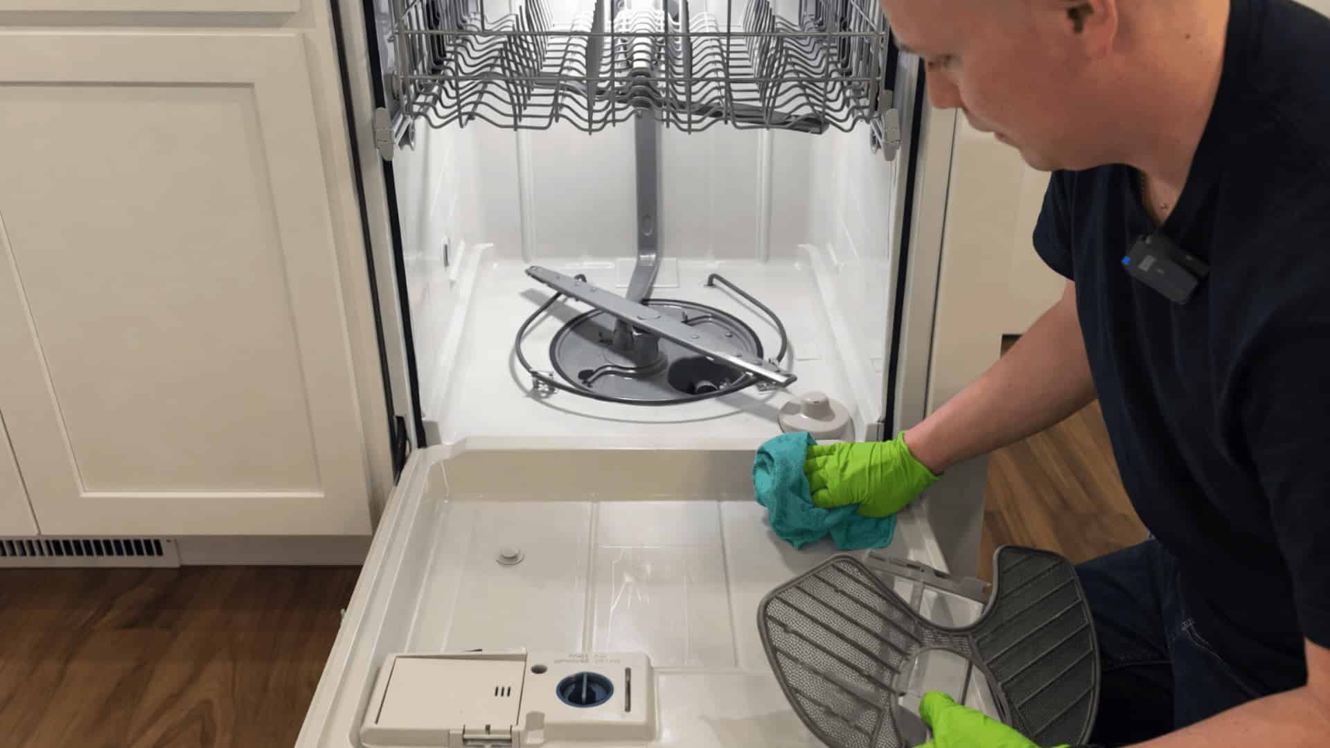 man wearing green gloves wiping inside of open dishwasher while holding removed filter screen