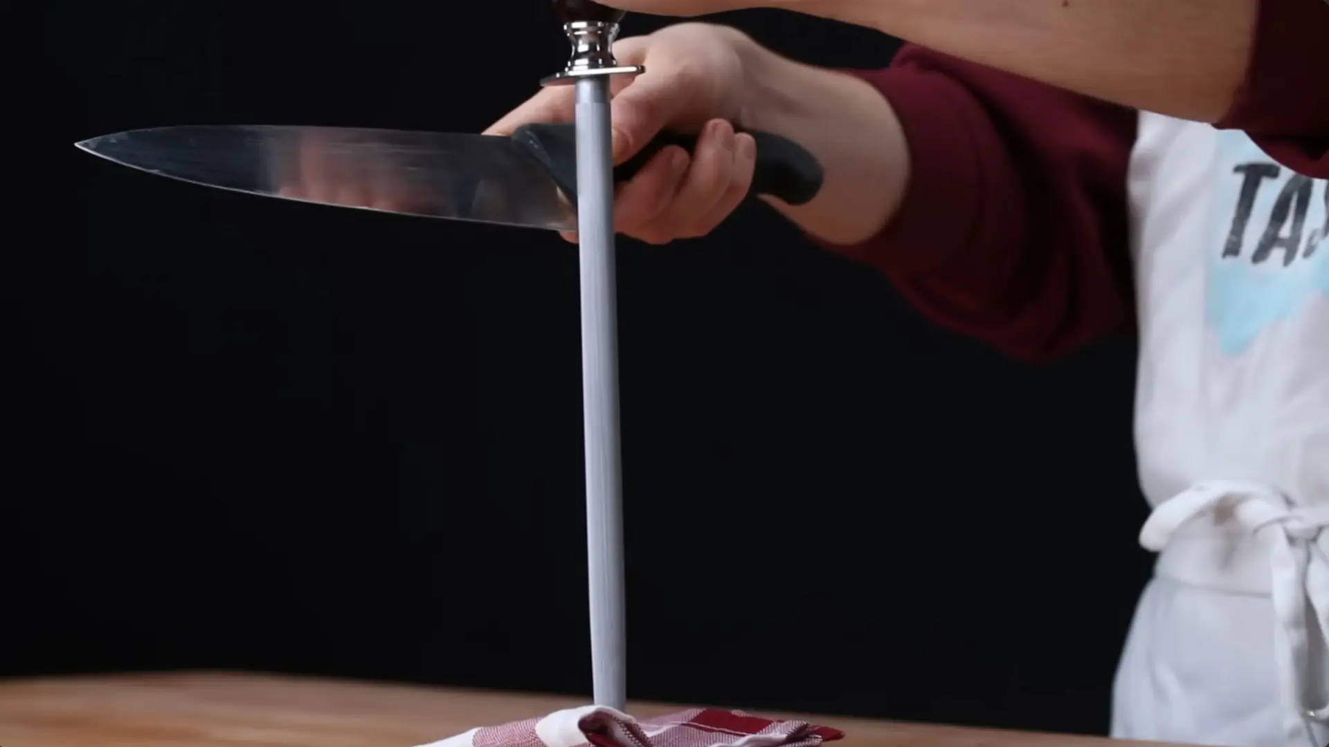 person uses a honing steel to realign the edge of a chef's knife over a wooden surface against a dark background