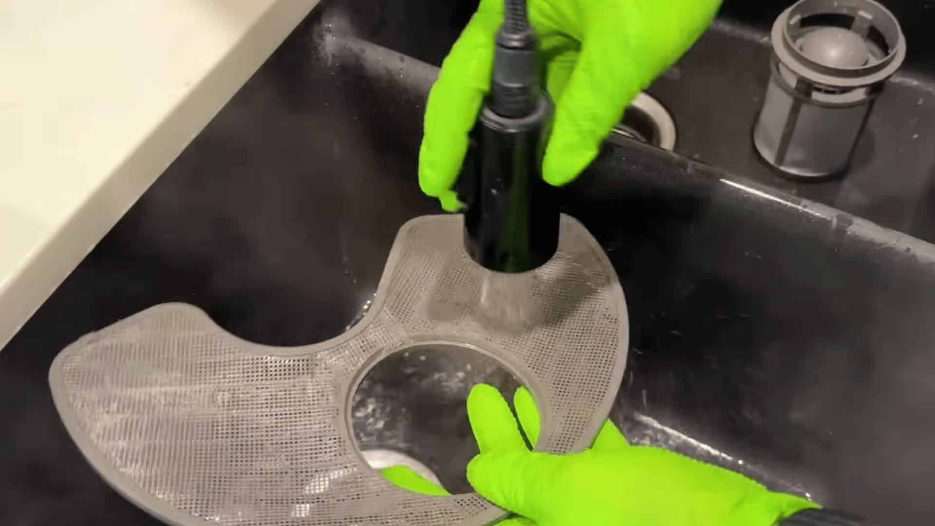 person wearing green gloves rinsing dishwasher filter under running water in kitchen sink