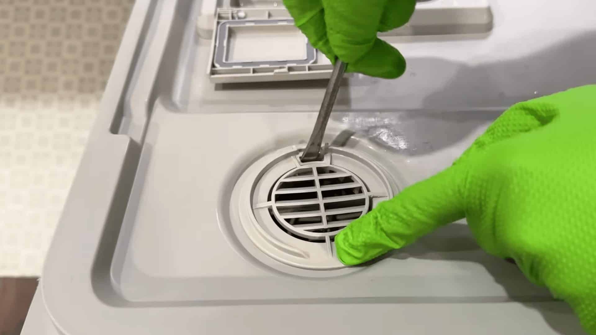 person wearing green gloves using screwdriver to remove dishwasher drain cover for cleaning