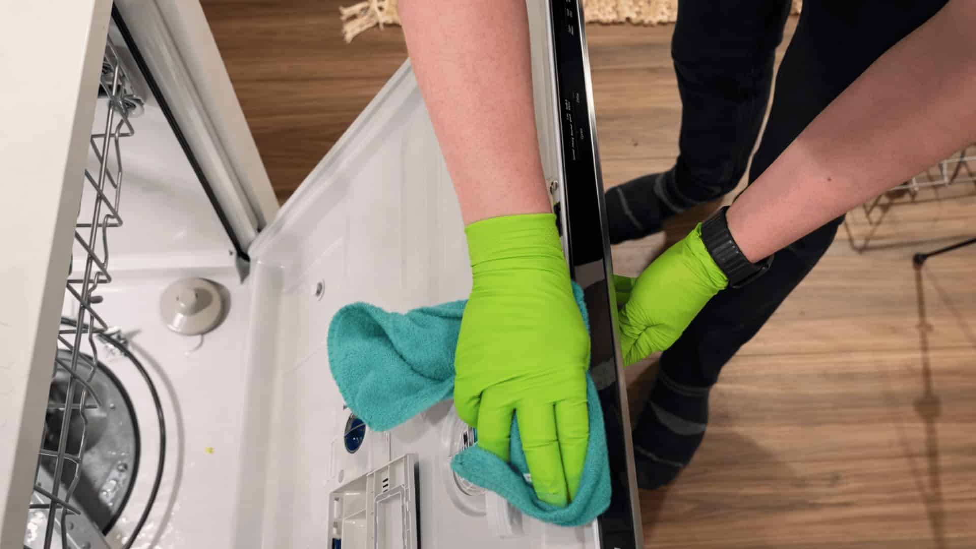 person wearing green gloves wiping inside edge of open dishwasher door with microfiber cloth