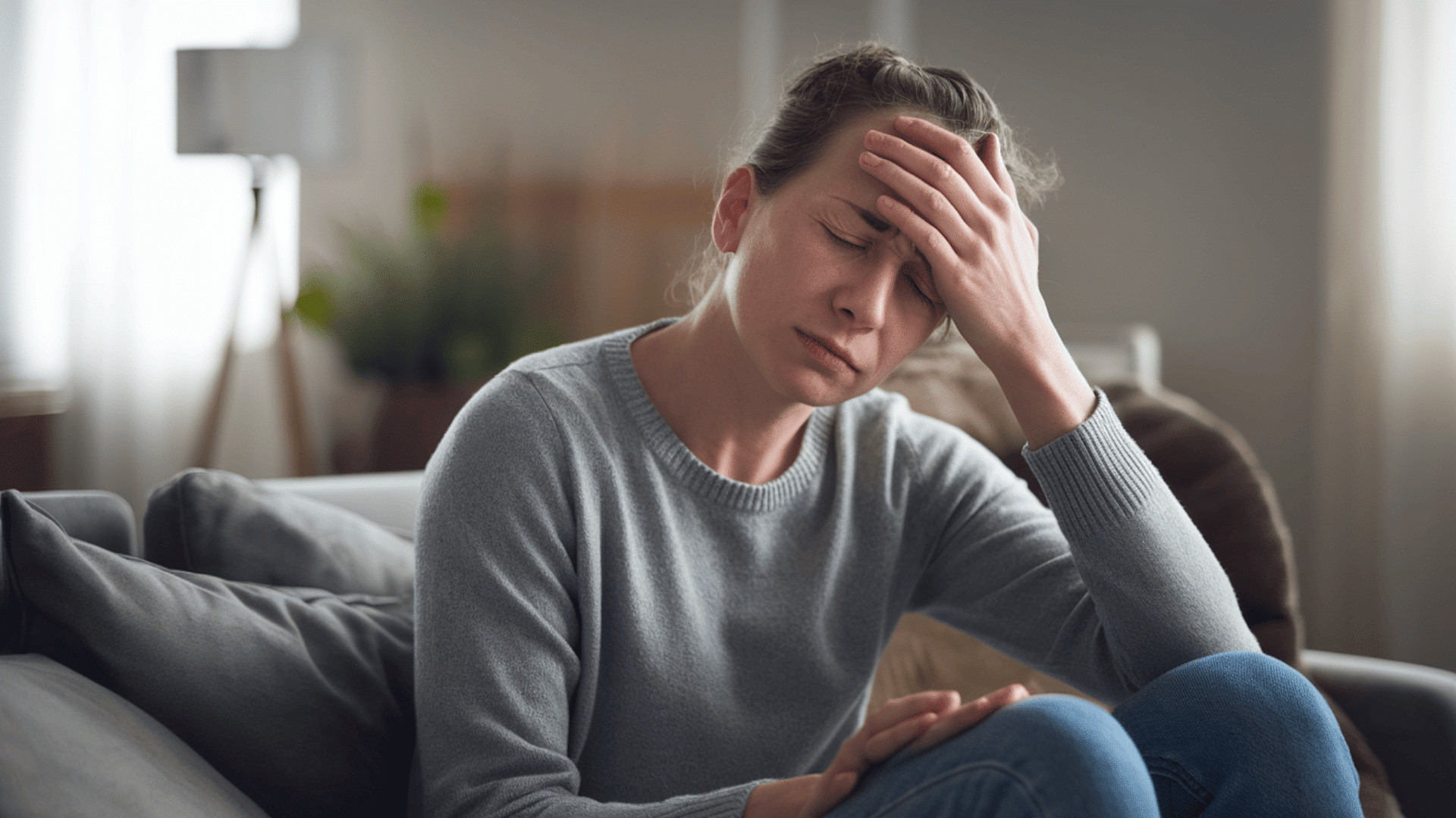 a woman sitting on a couch with a hand on her forehead, appearing to be in pain or discomfort.