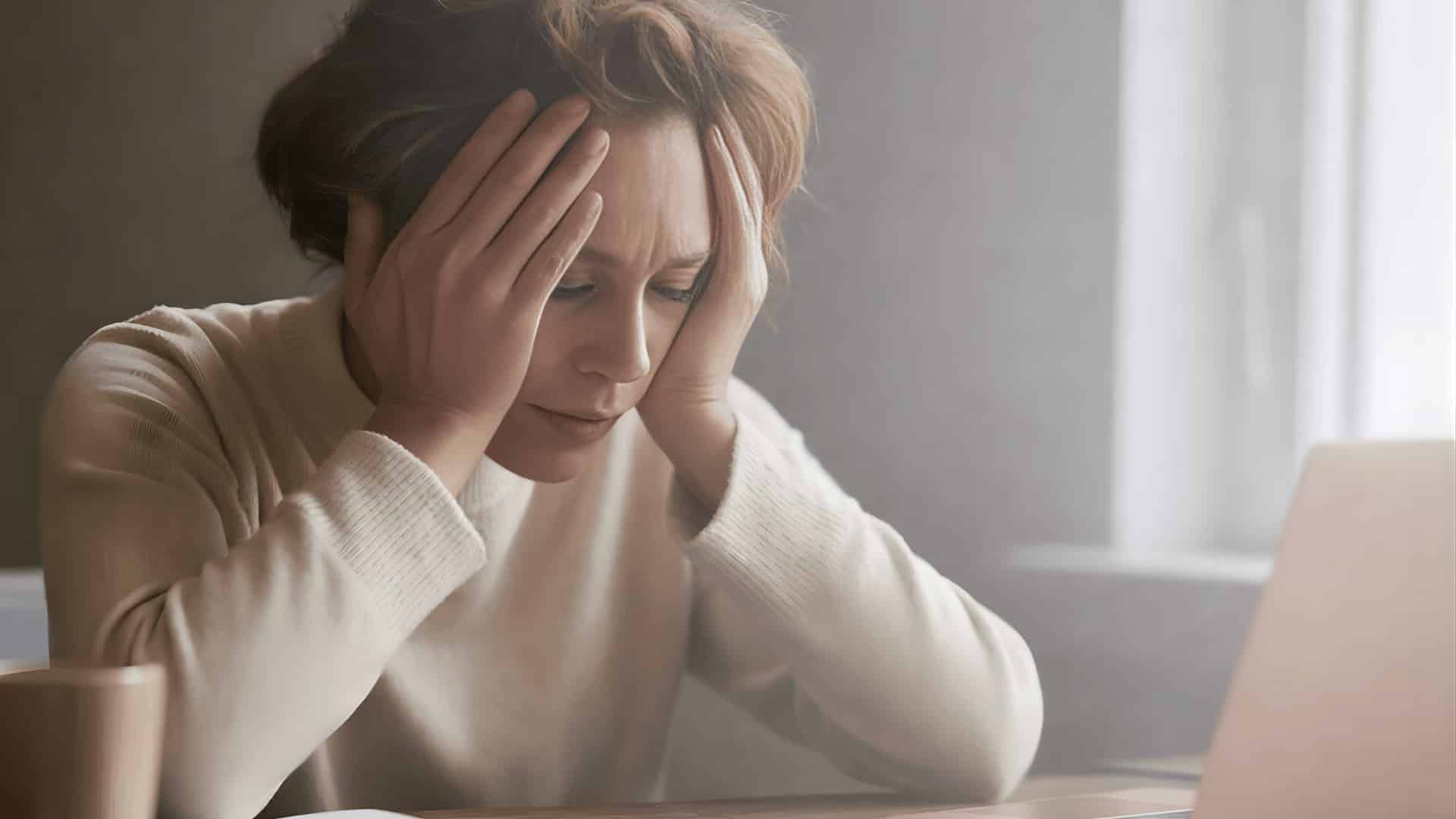 woman sitting at a desk, looking stressed with her hands on her head, laptop and coffee cup in the background (1)