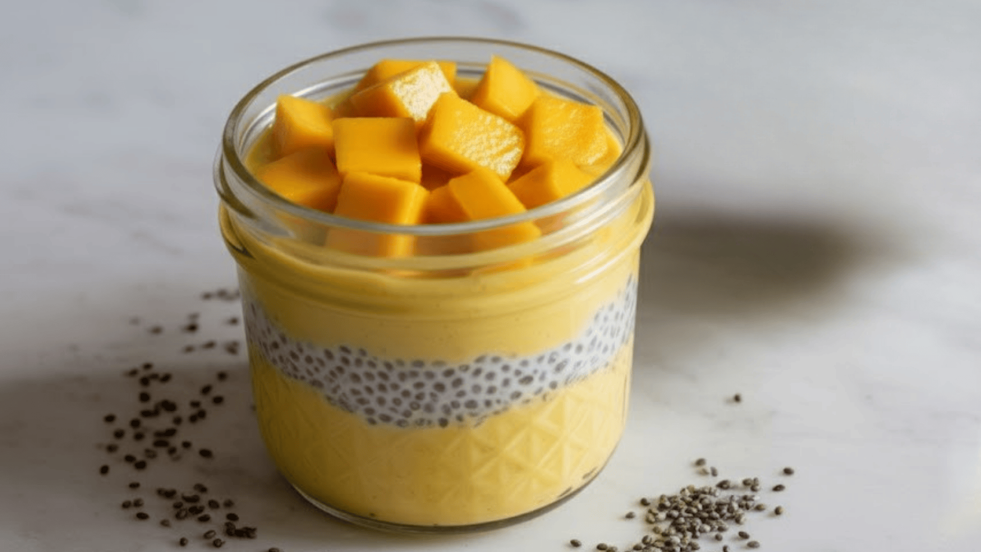 a mason jar with mango chia pudding topped with fresh diced mango chunks, with a cup of green tea on a white surface