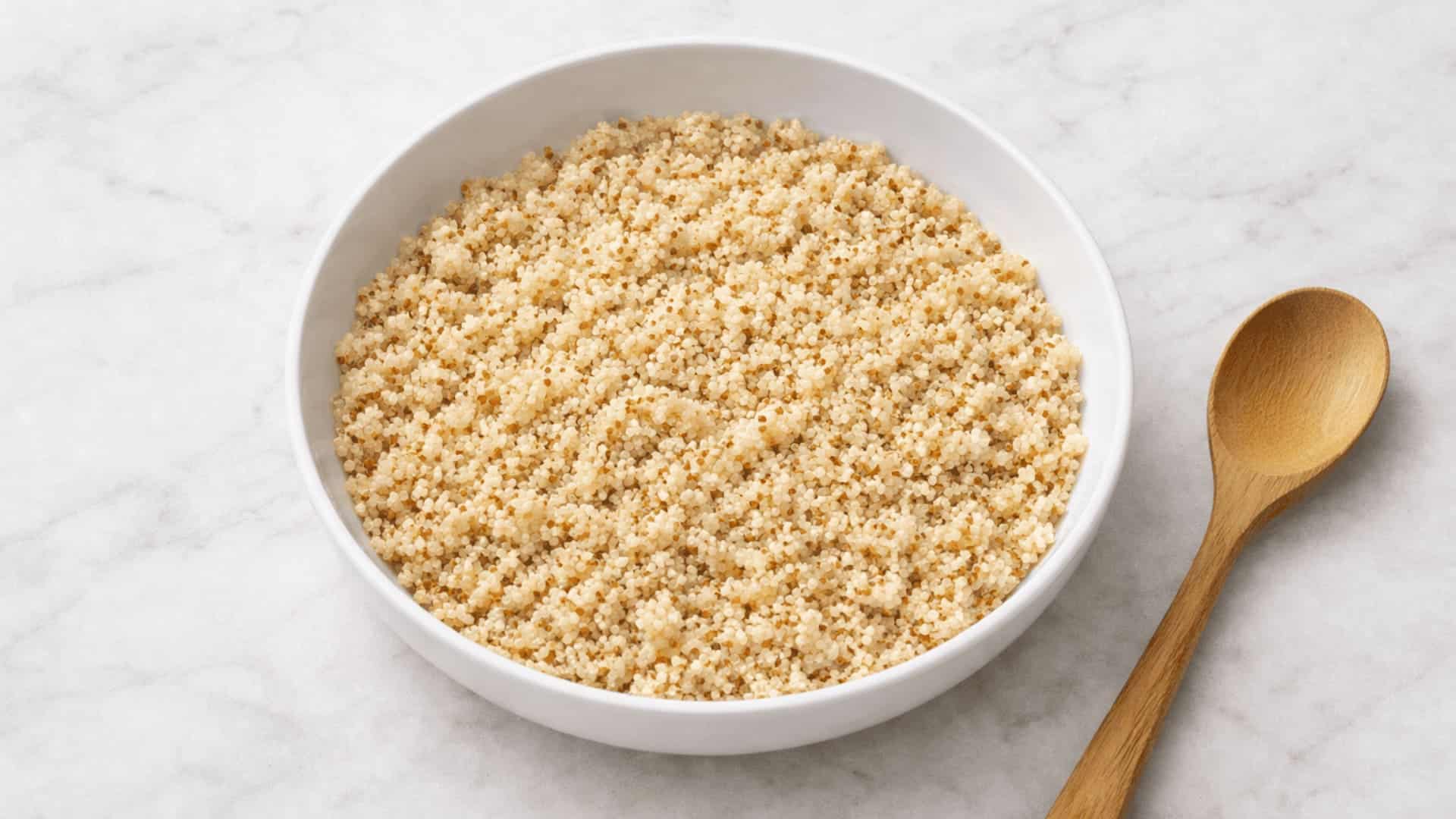 a white bowl of fluffy cooked quinoa with a wooden spoon on the side placed on a marble surface