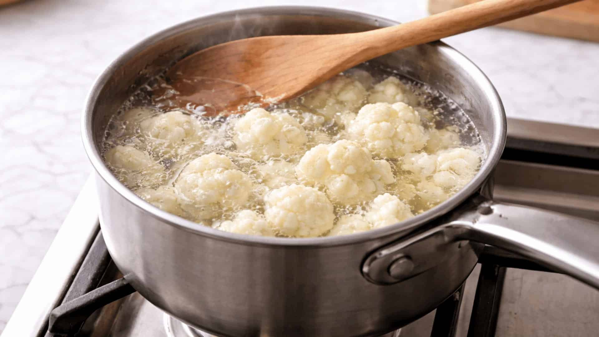 cauliflower florets boiling in a stainless steel saucepan on a gas stove with a wooden spoon resting on top