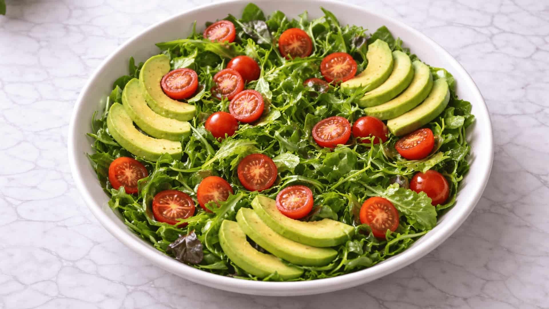 fresh mixed greens salad with sliced avocado and halved cherry tomatoes in a white bowl on a marble surface