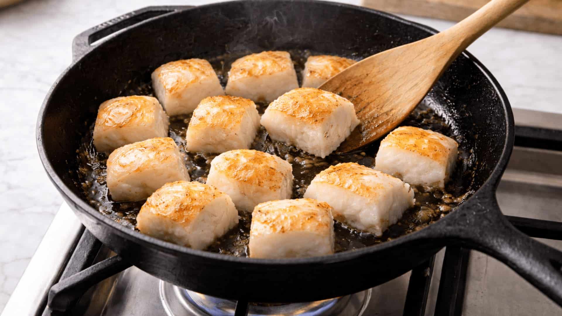 golden-seared cubed white fish fillets cooking in a cast iron skillet on gas stove with wooden spatula
