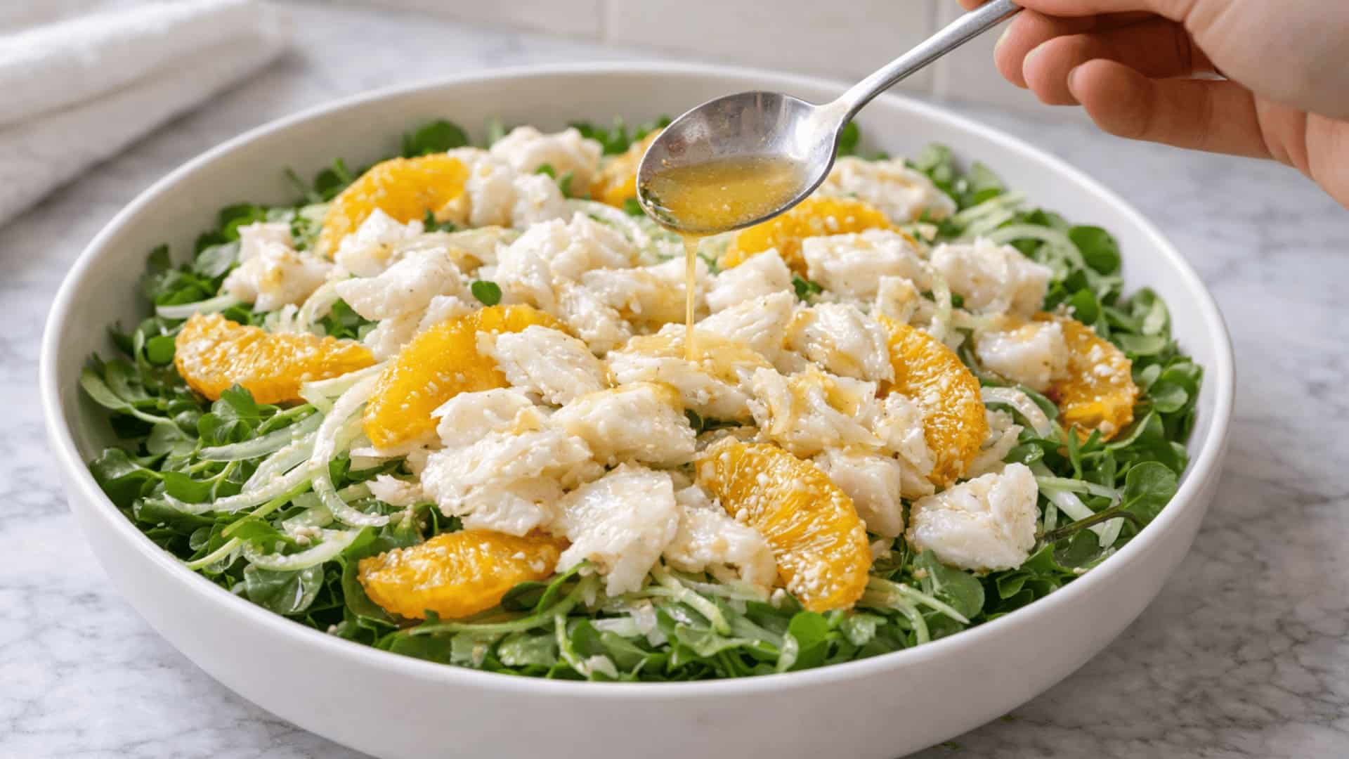hand drizzling white wine vinaigrette over poached cod salad with fennel, orange segments, and watercress