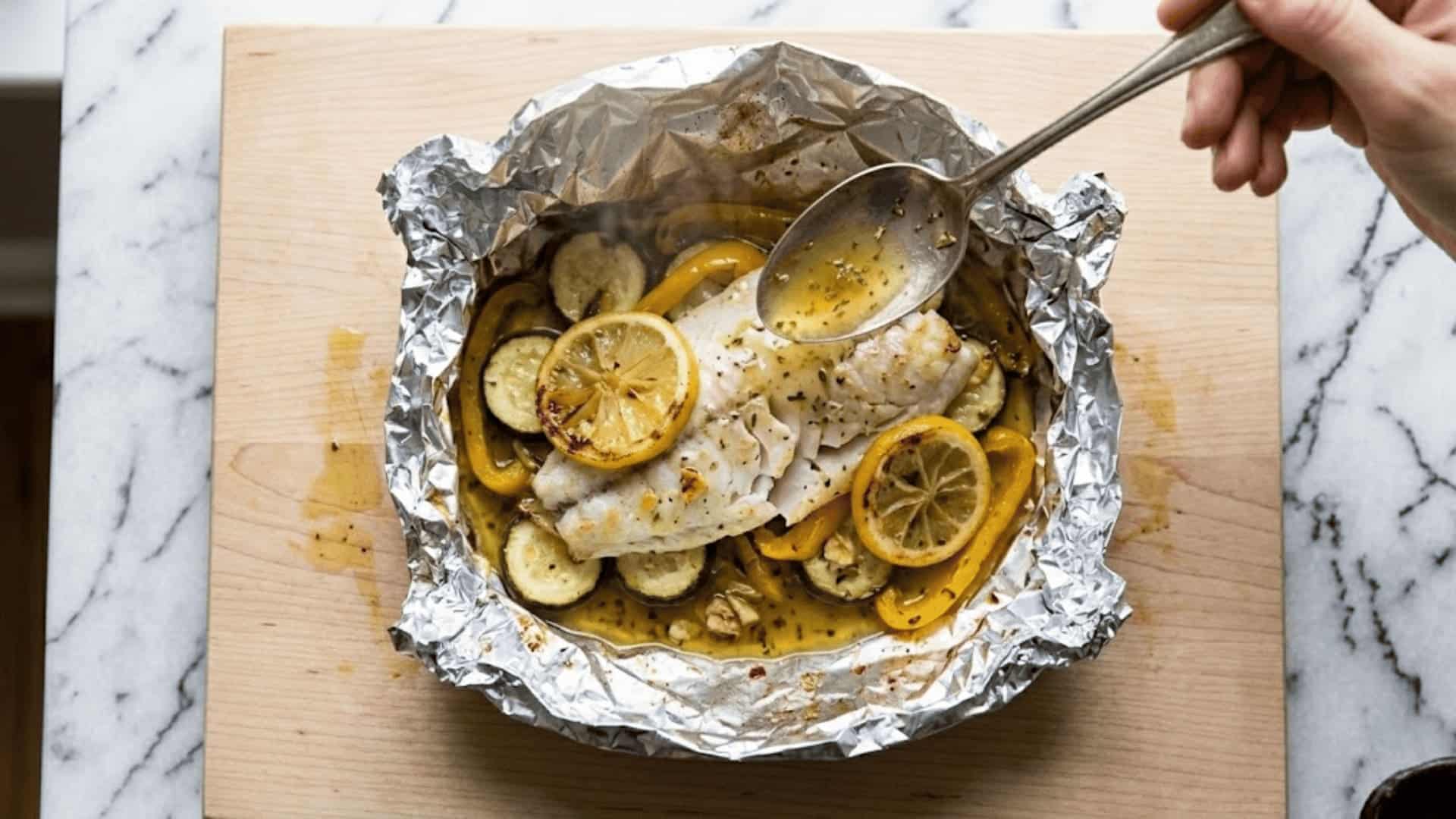 hand spooning juices over a cooked fish fillet with zucchini, bell pepper, and lemon in an open foil packet