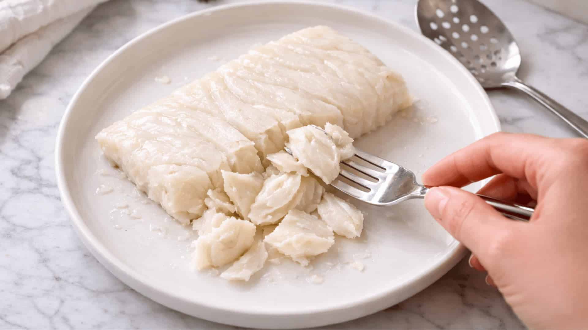 hand using a fork to flake poached cod fillet into tender pieces on a white plate over marble surface
