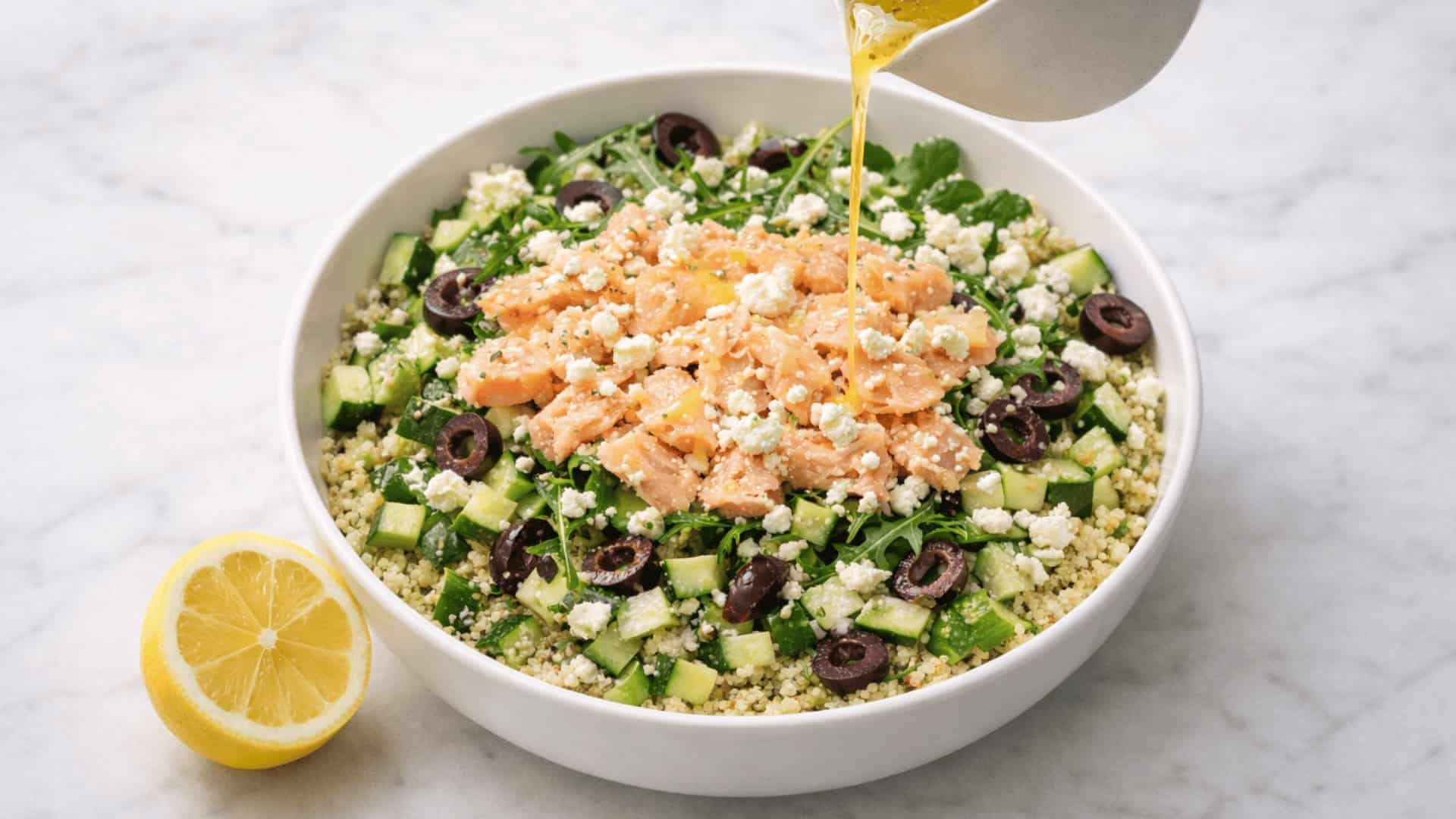 lemon dressing being poured over a quinoa salmon bowl with cucumber, black olives, feta, and arugula on marble
