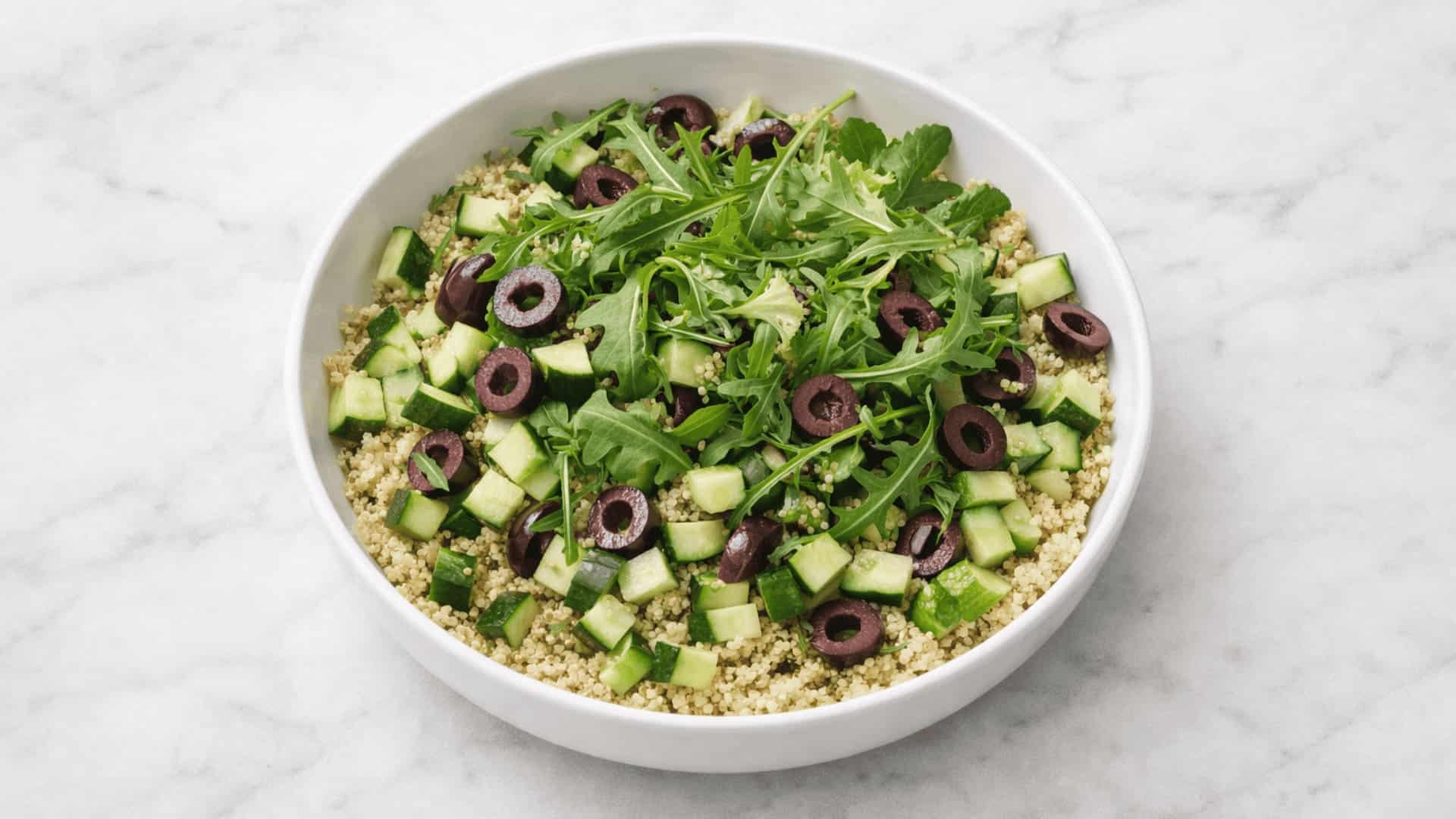 quinoa salad bowl with diced cucumber, sliced black olives, and fresh arugula in a white bowl on marble
