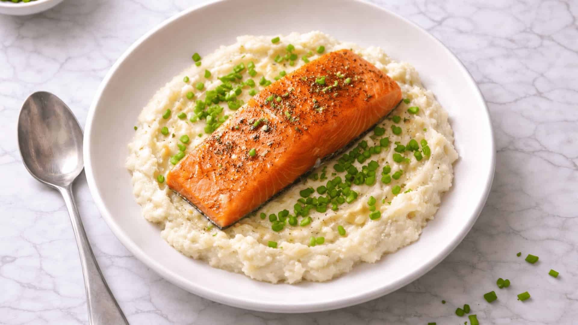 roasted salmon fillet on creamy cauliflower mash topped with chopped chives in a white bowl on a marble surface