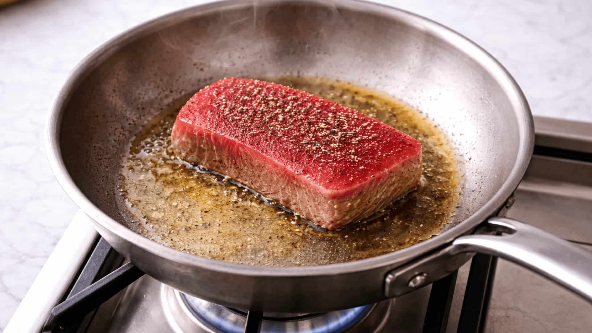 seasoned tuna steak searing in a stainless steel pan with hot oil over a blue gas flame on a stovetop