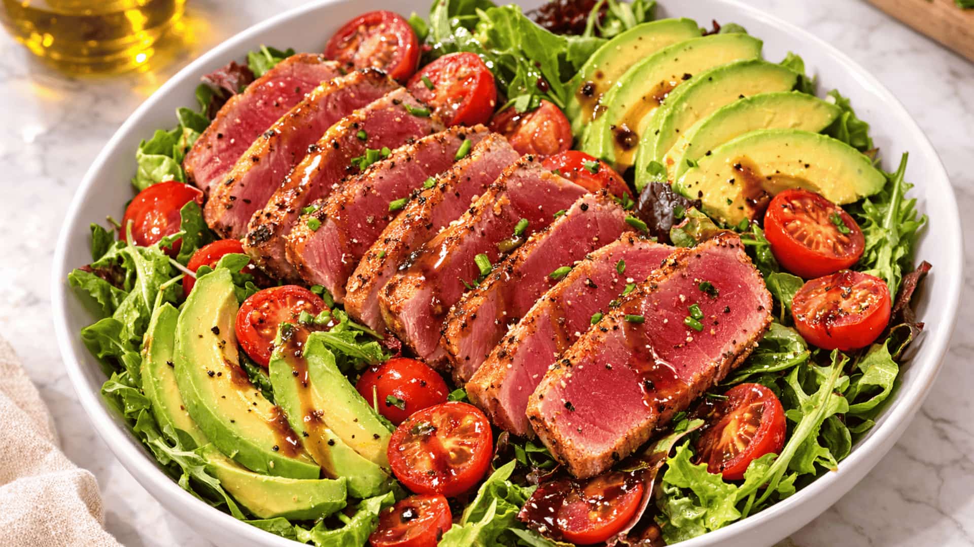 sliced seared tuna steak over mixed greens with avocado, cherry tomatoes, and dark dressing in a white bowl