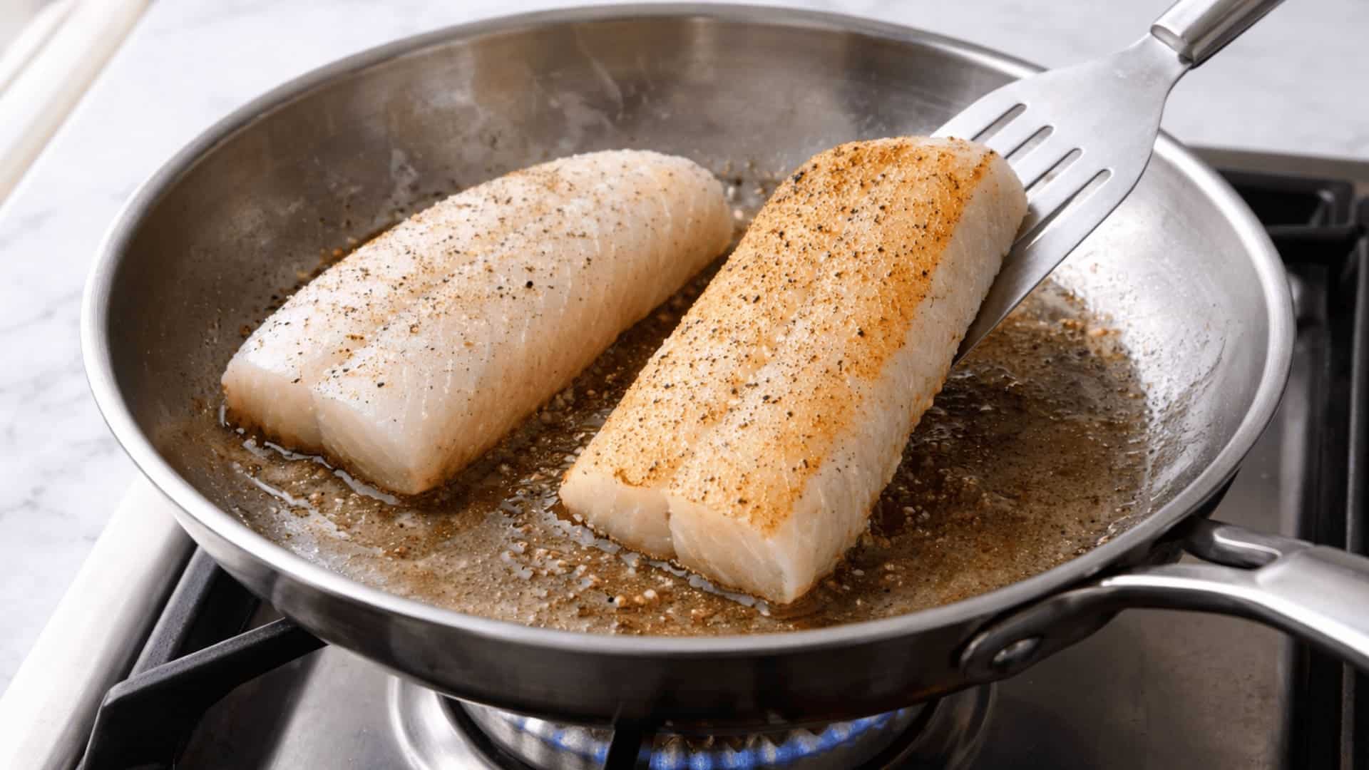 two cod fillets pan searing in a stainless steel skillet over a gas stove with a golden brown crust