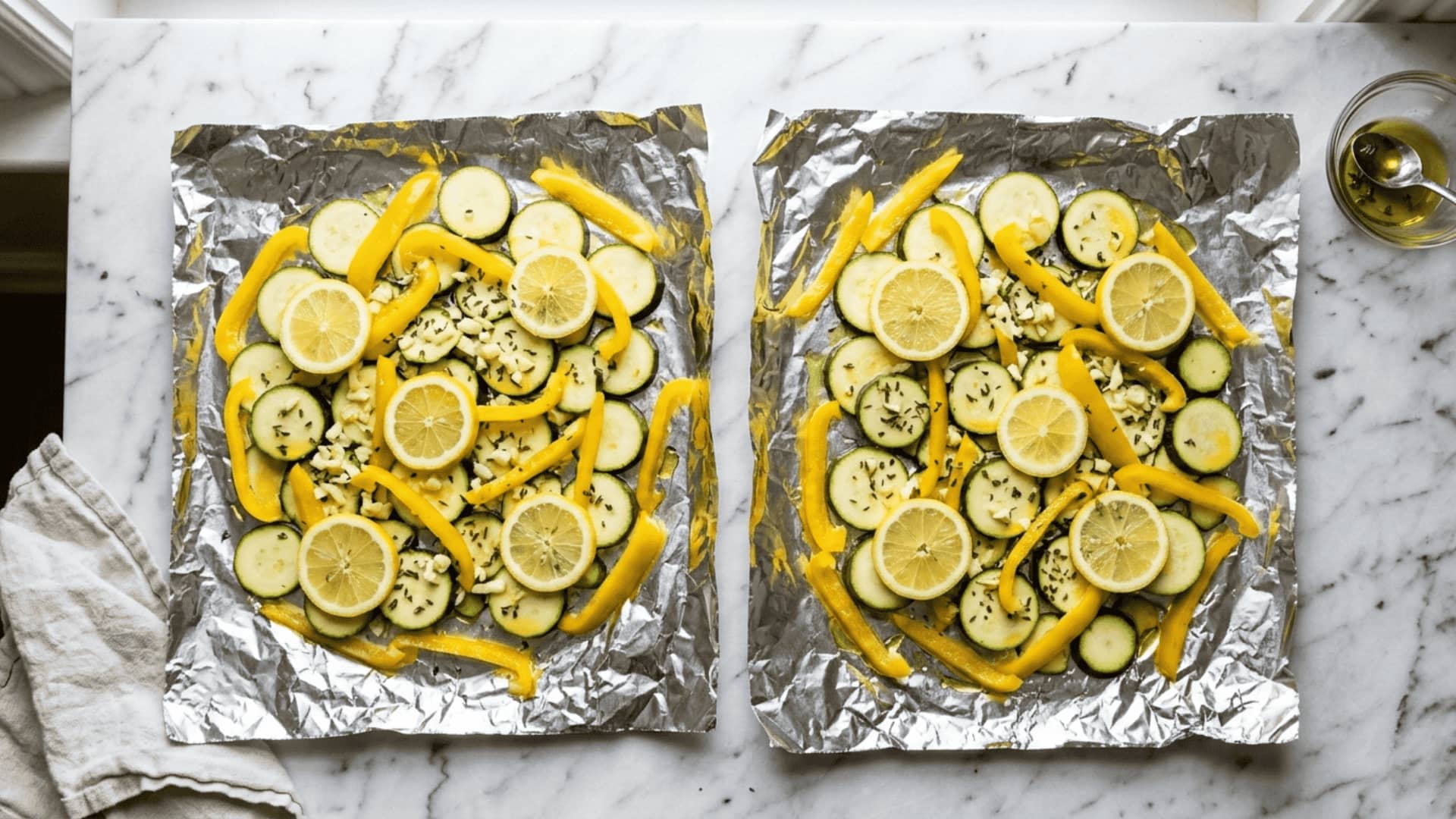 two open foil packets filled with sliced zucchini, yellow bell pepper, lemon rounds, and minced garlic on marble