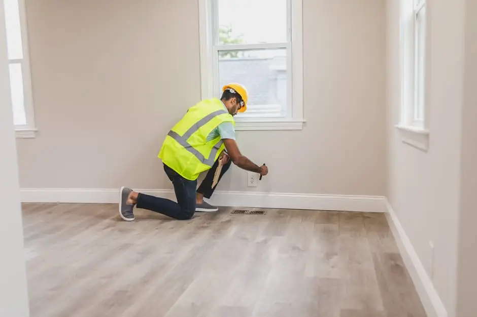 Worker in safety gear inspecting electrical outlet in empty room with hardwood floor
