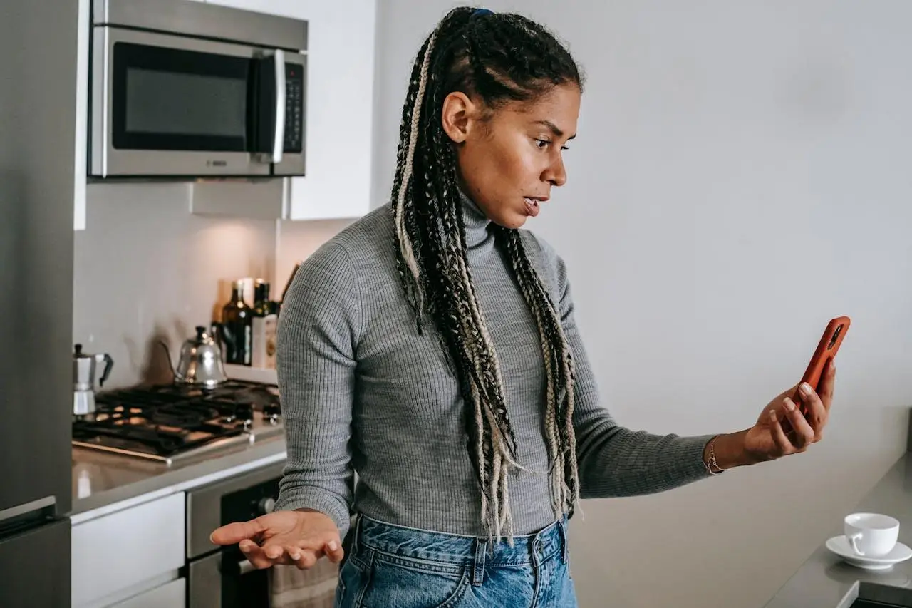 Person in gray sweater holding smartphone in modern kitchen with stainless steel appliances