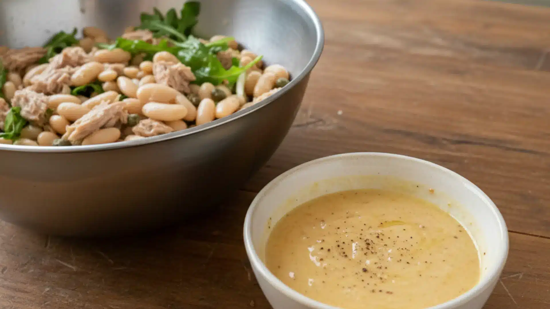 creamy yellow dressing in a small white bowl next to a large metal bowl of tuna and bean salad on a wooden table