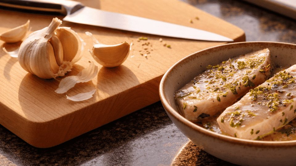 raw swordfish steaks soaking in lemon herb marinade in a ceramic dish with a garlic bulb and cloves on a wooden board