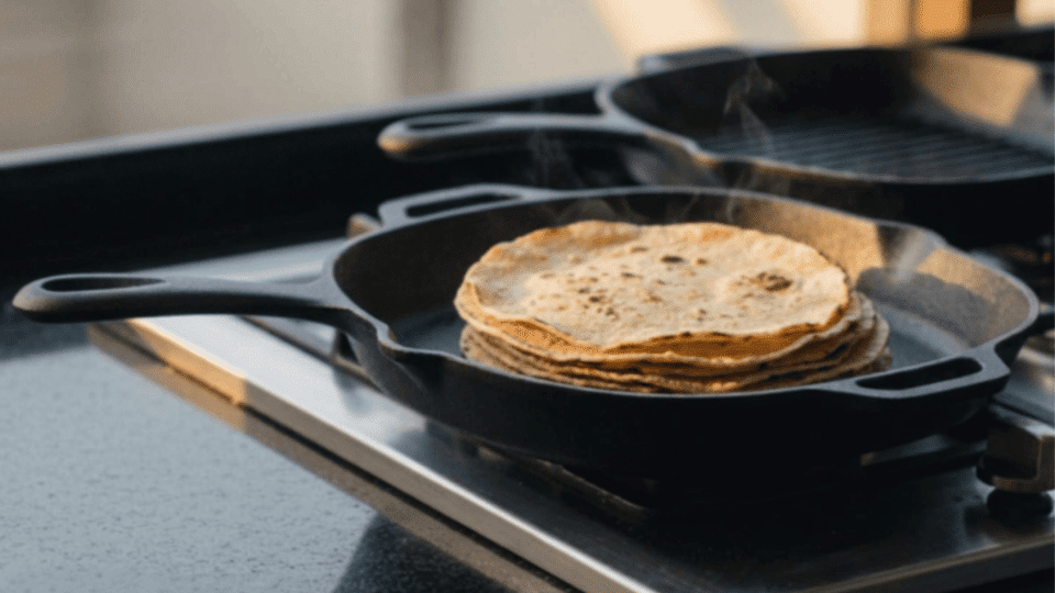 stack of warm corn tortillas heating in a cast iron skillet on a gas stove with a small bowl of salt nearby