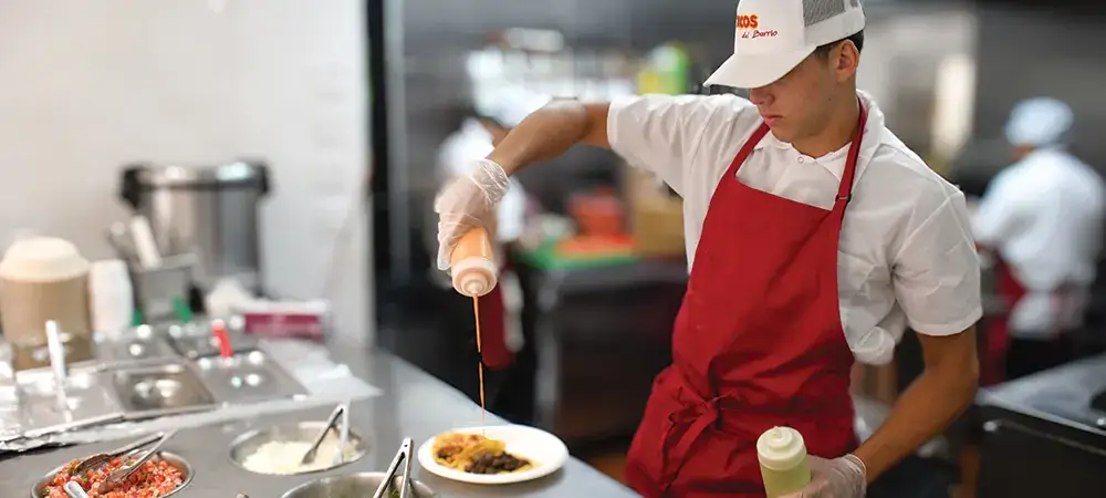 Worker in red apron drizzling sauce over taco in restaurant kitchen setting