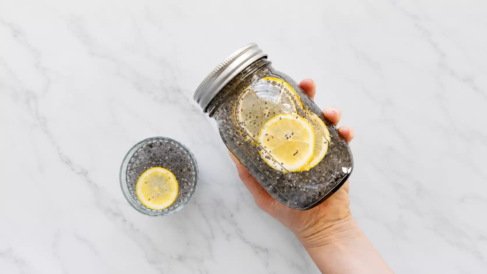 Hand holding a mason jar of lemon chia water beside a glass with lemon slices on a marble countertop