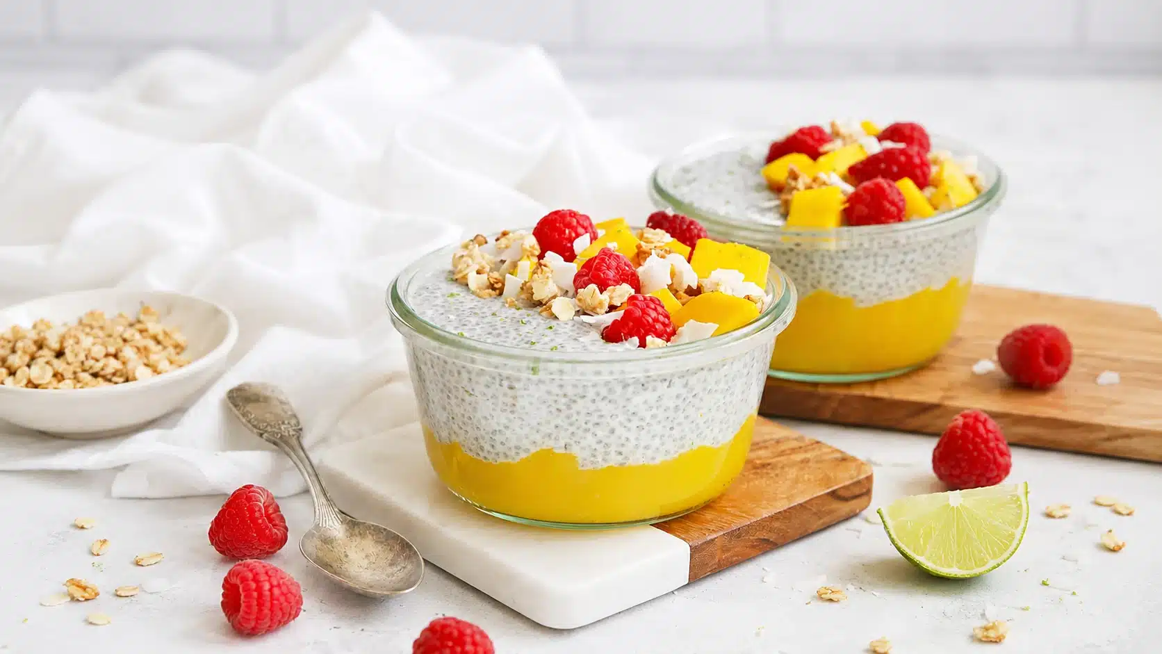 Layered mango chia pudding bowls topped with raspberries, mango chunks, coconut flakes, and granola on a bright table