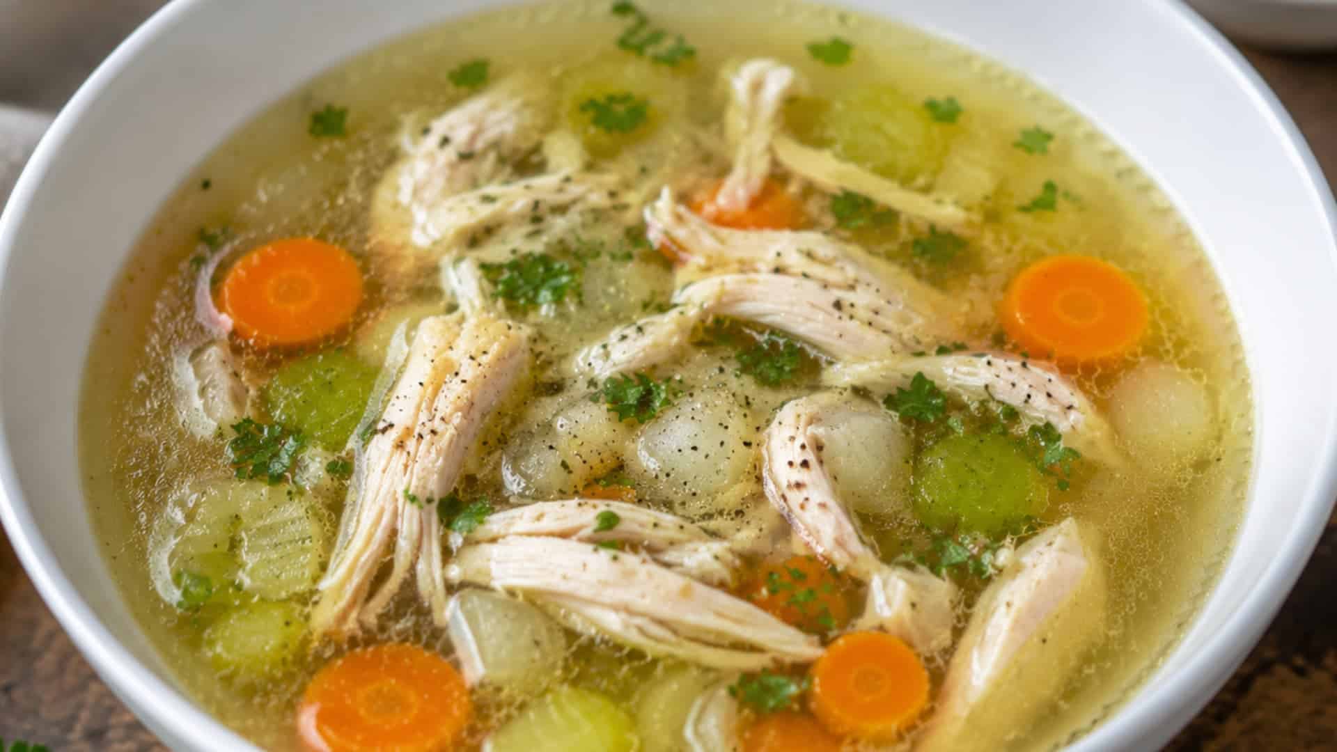 bowl of clear chicken soup with shredded chicken carrots celery and fresh herbs on wooden surface
