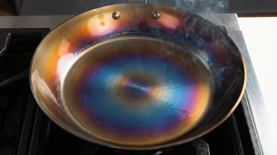 carbon steel pan developing a rainbow patina with visible smoke while being heated on a stovetop as part of the seasoning process