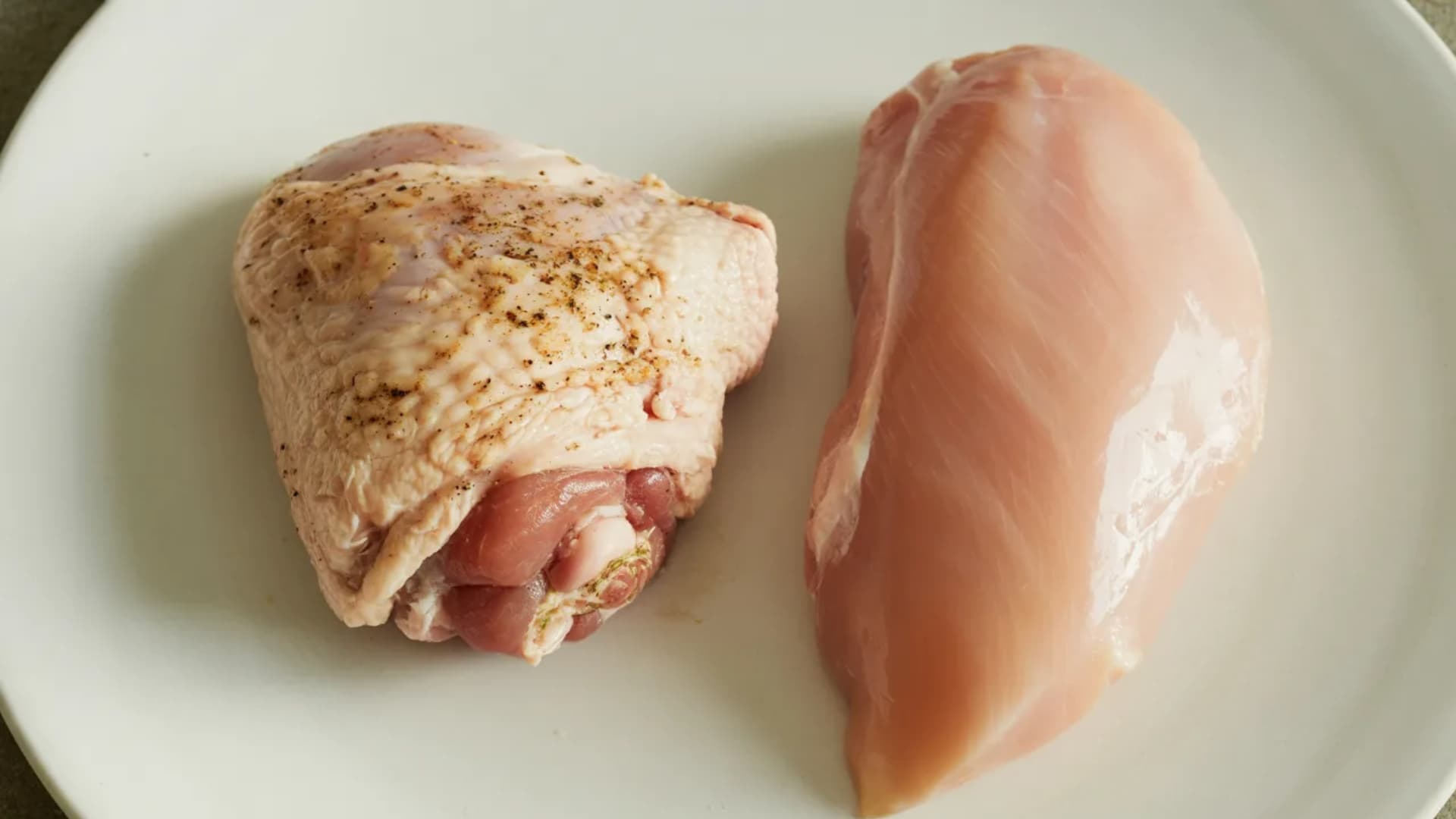 chicken thigh and chicken breast side by side on plate, clear difference in texture and size, minimal background, realistic food photography