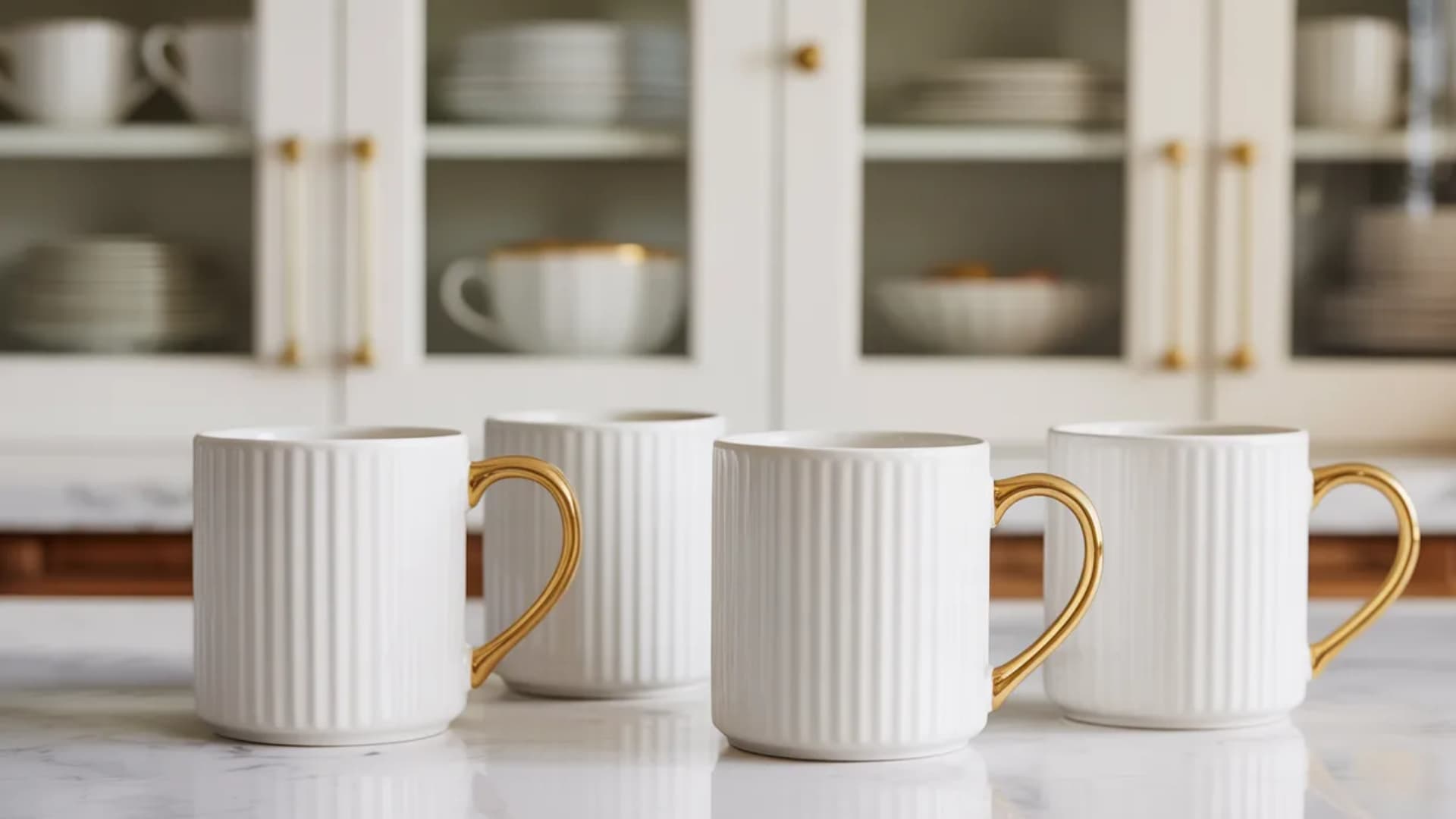 close-up of white ceramic mugs with gold handles arranged on a marble countertop. The mugs feature a textured ribbed design, and the gold handles add an elegant touch to the clean, modern aestheti