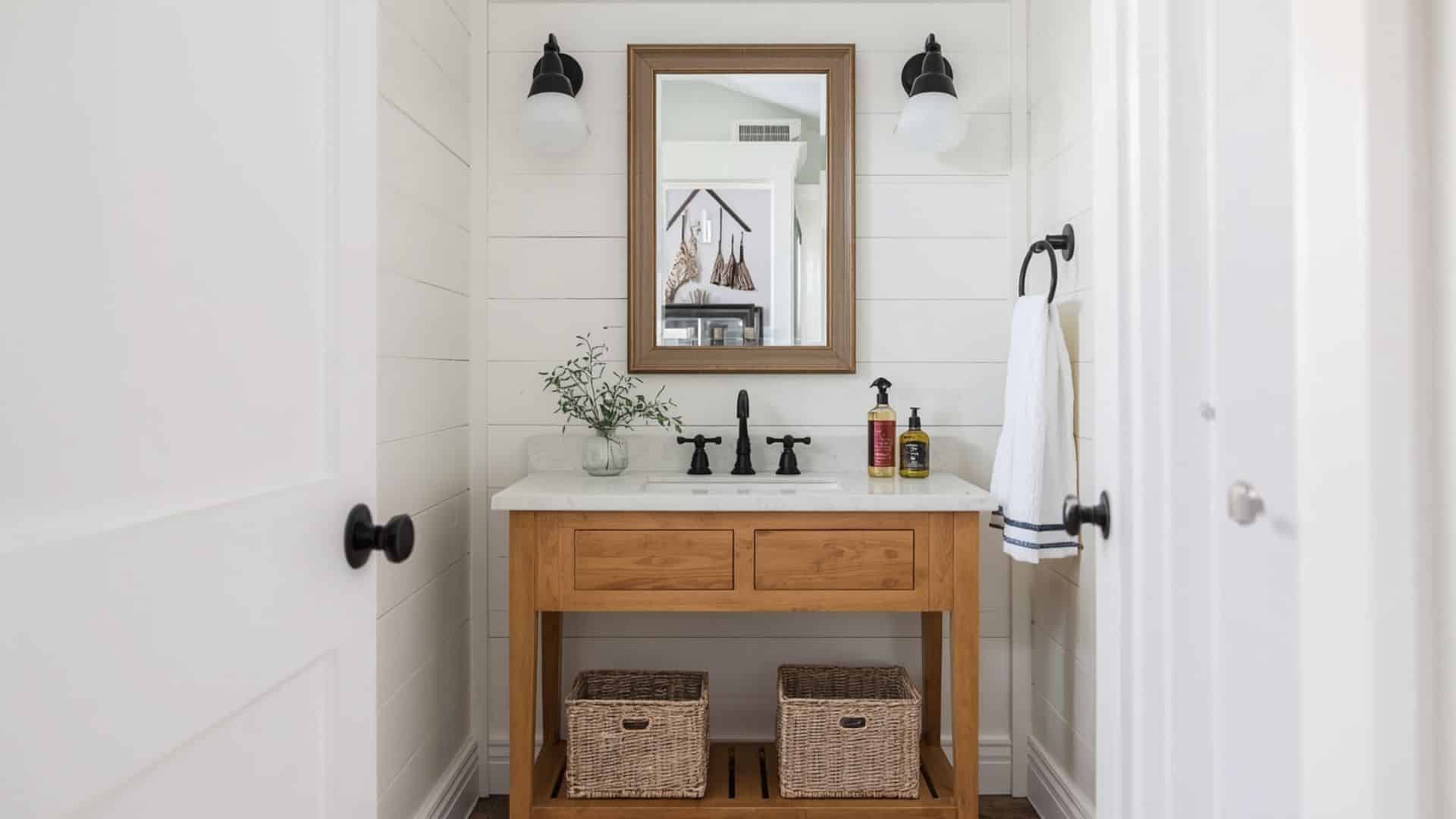 farmhouse bathroom with white shiplap walls, warm wood vanity, vintage-style mirror, black or chrome fixtures, woven baskets, light neutral palette, cozy clean design