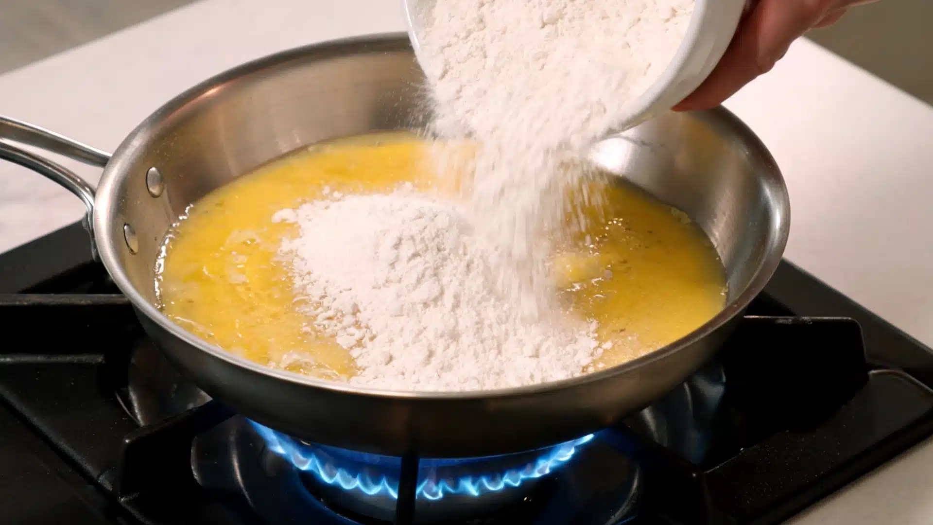 flour being poured into melted butter in a pan on a gas stove to start making roux for sauce base